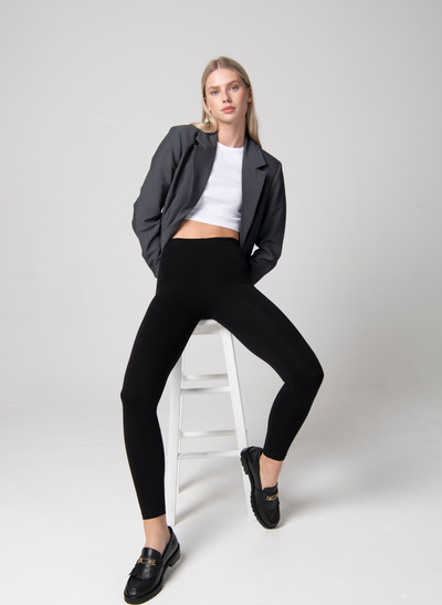 Woman wears Black Leggings, White Top, Grey Blazer and Black Brogue shoes. She sits on a white stool in a studio room.
