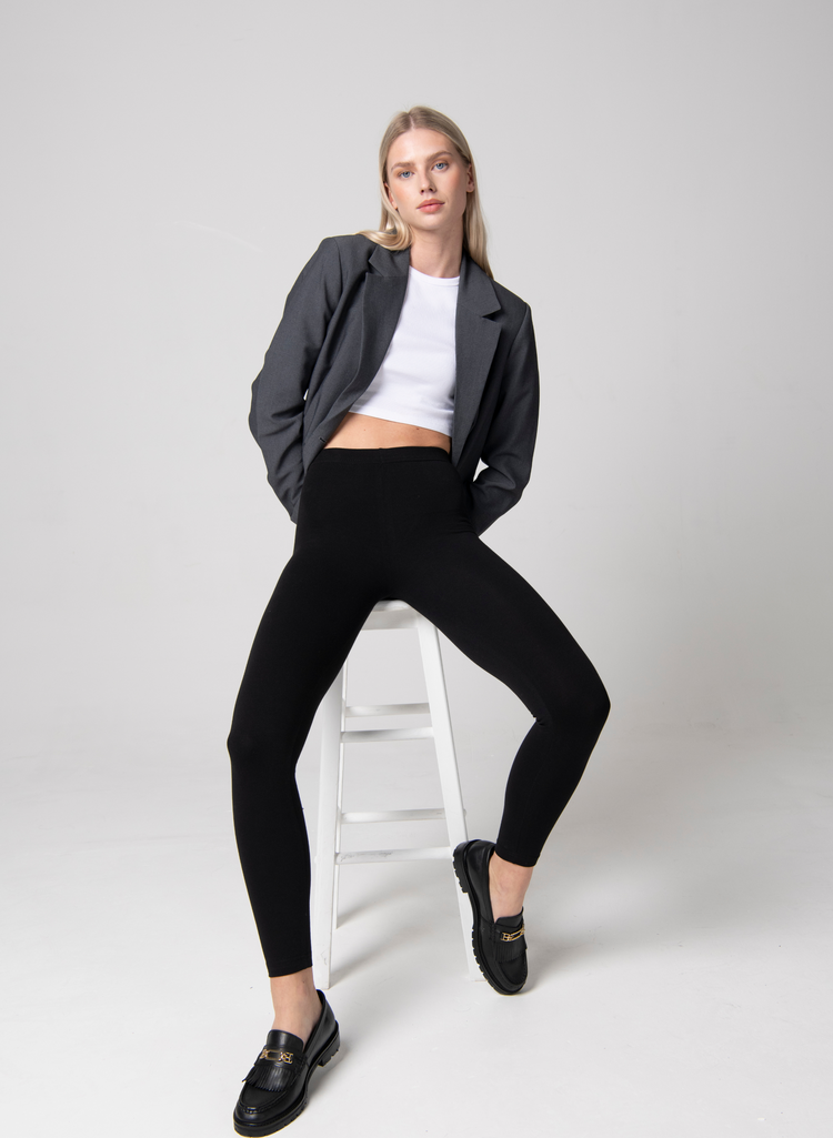 Woman wears Black Leggings, White Top, Grey Blazer and Black Brogue shoes. She sits on a white stool in a studio room.